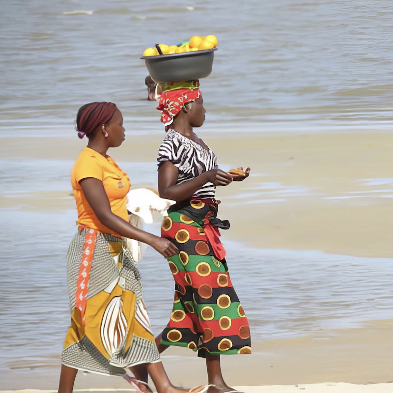 Two colourfully dressed women walk along a sandy beach. One has a basket of fruit on her head.